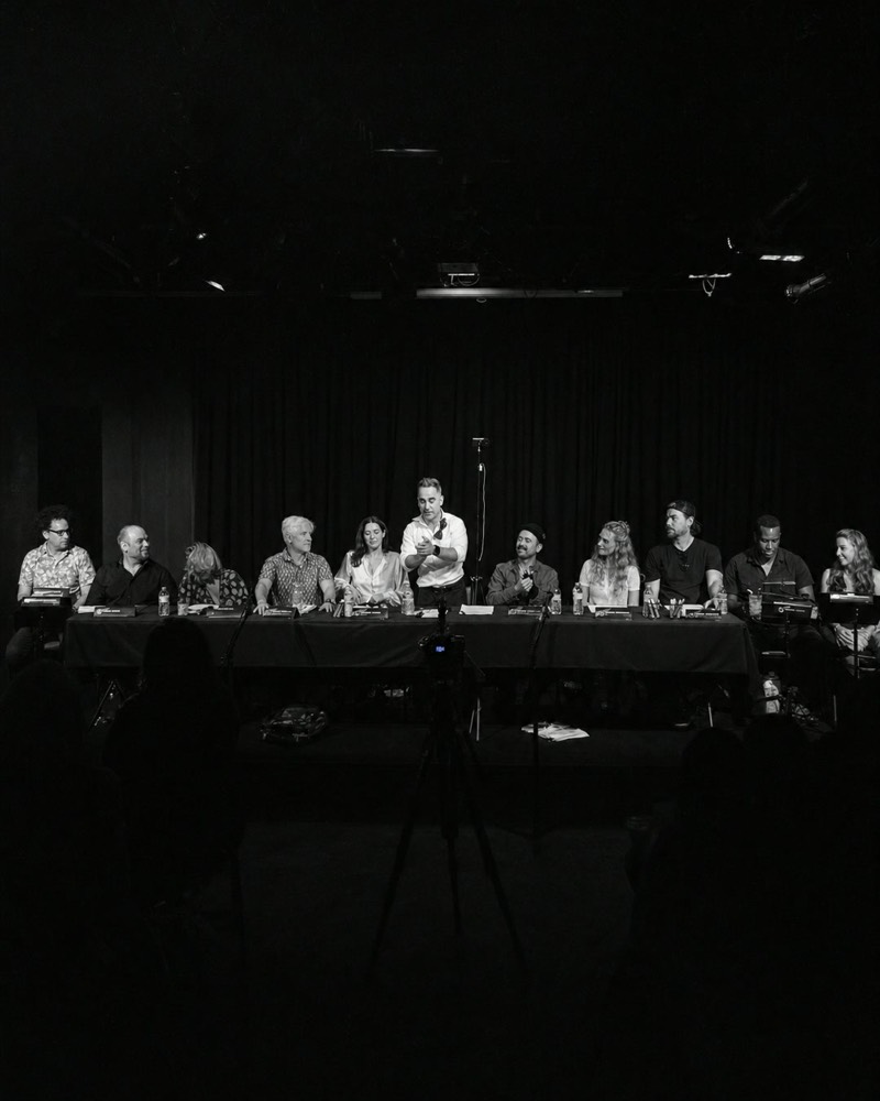 The full cast seated onstage at the table read